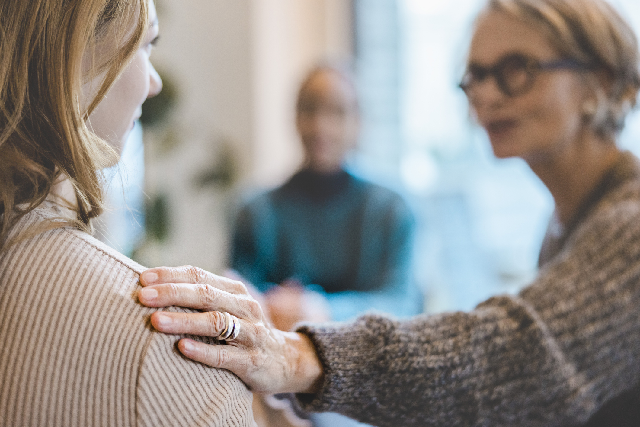 A person with their back to the camera has a person sat across from them extending their hand out to the person's shoulder, someone is blurred in the background sat at the same table.