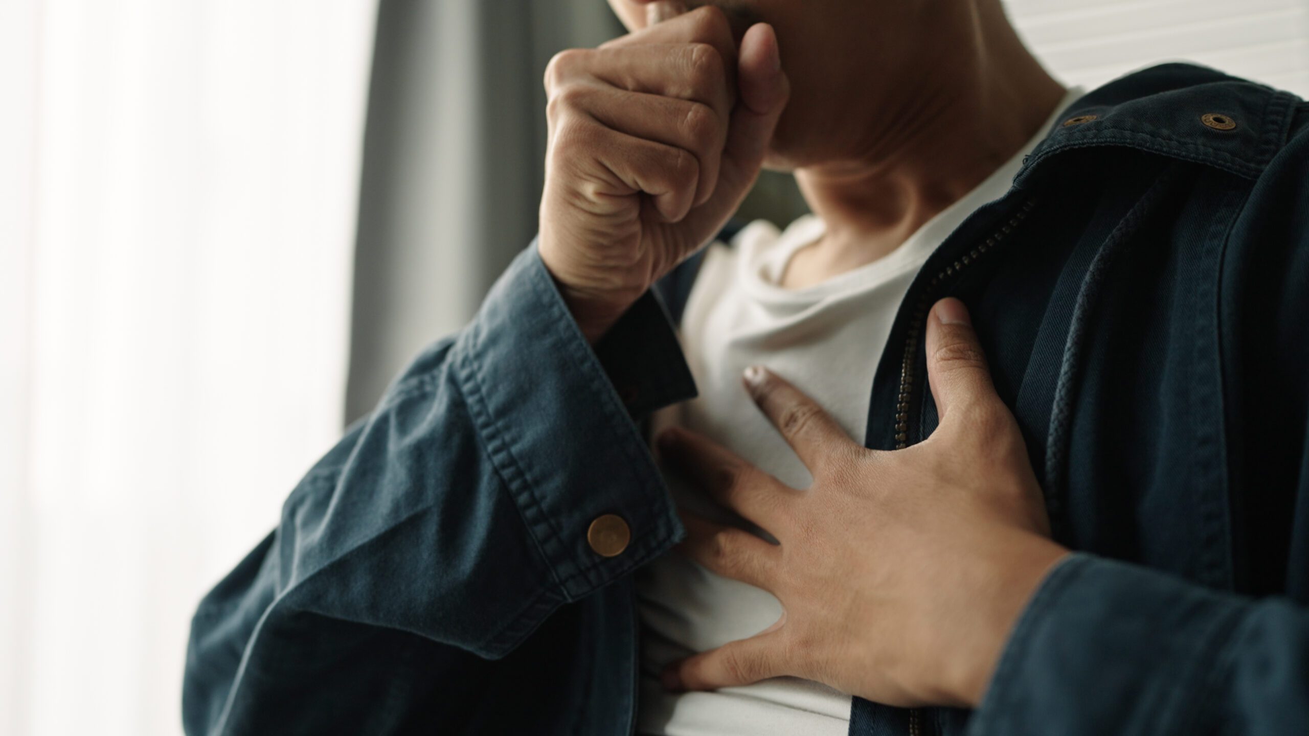 Close up of a person covering their mouth with one hand as they cough, their other hand is laid on their chest