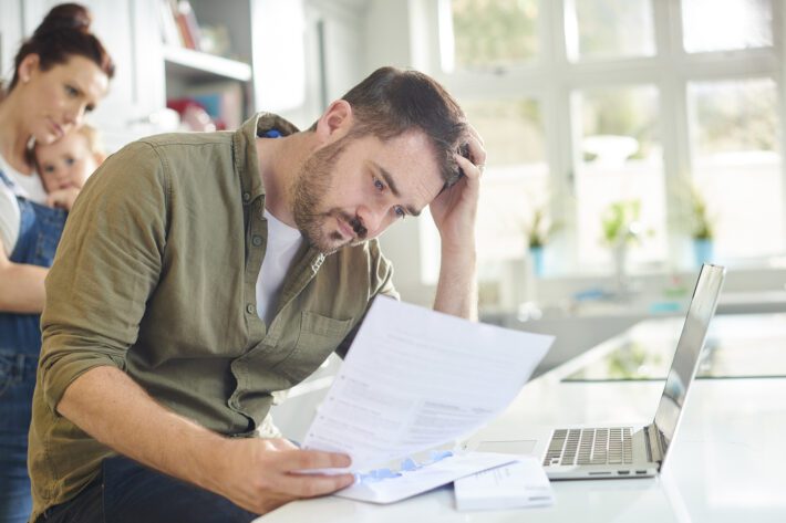 A man sits with his head in his hand at his laptop holding a piece of paper looking worried, his partner stands behind him holding a baby.