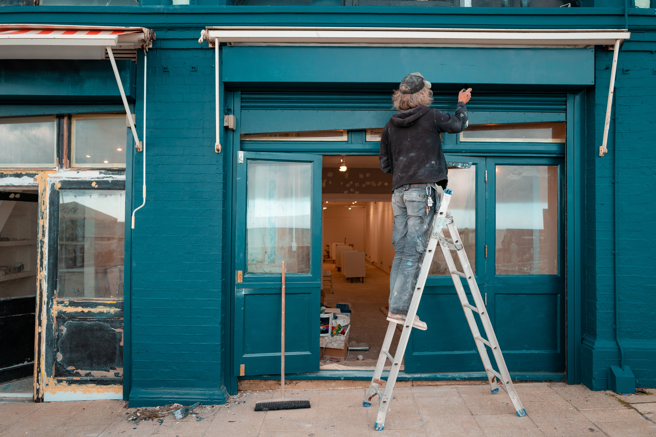 A workman stands on a ladder outside of a shop painting, we can see the inside is being refurbished.