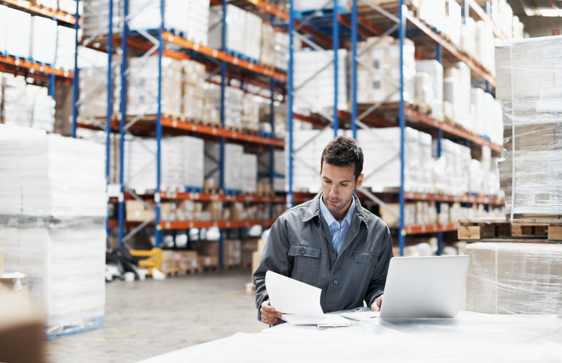 A manager sits in a warehouse at his laptop looking over pieces of paper.