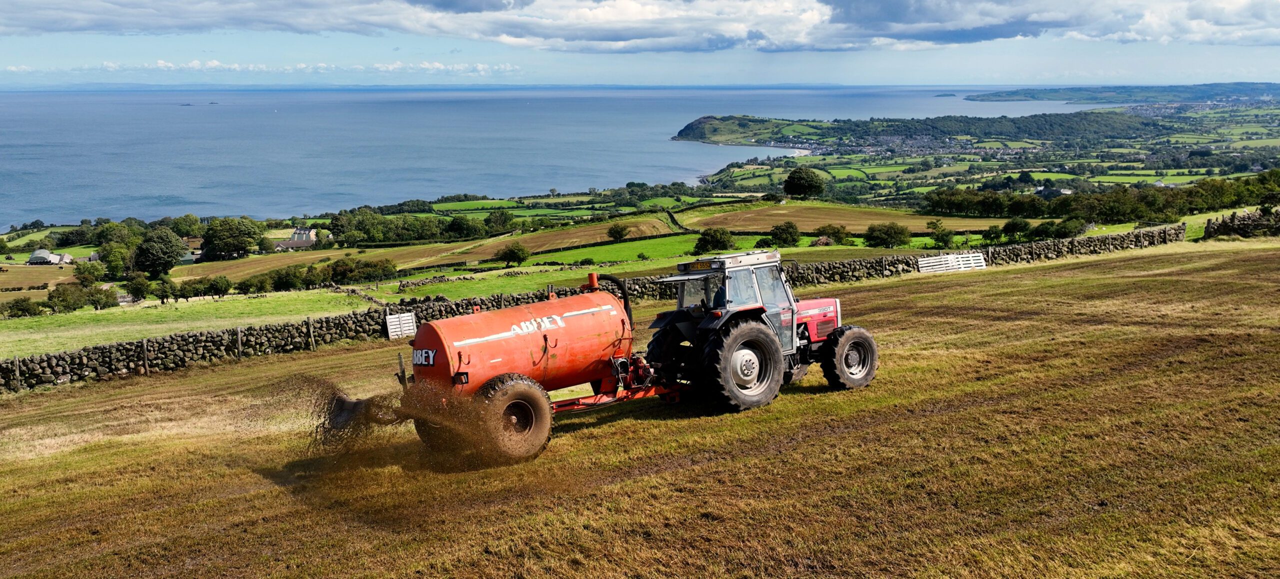 An aerial shot of a tractor with a tanker full of slurry attached being dispersed out onto some fields, the sea can be seen in the distance.