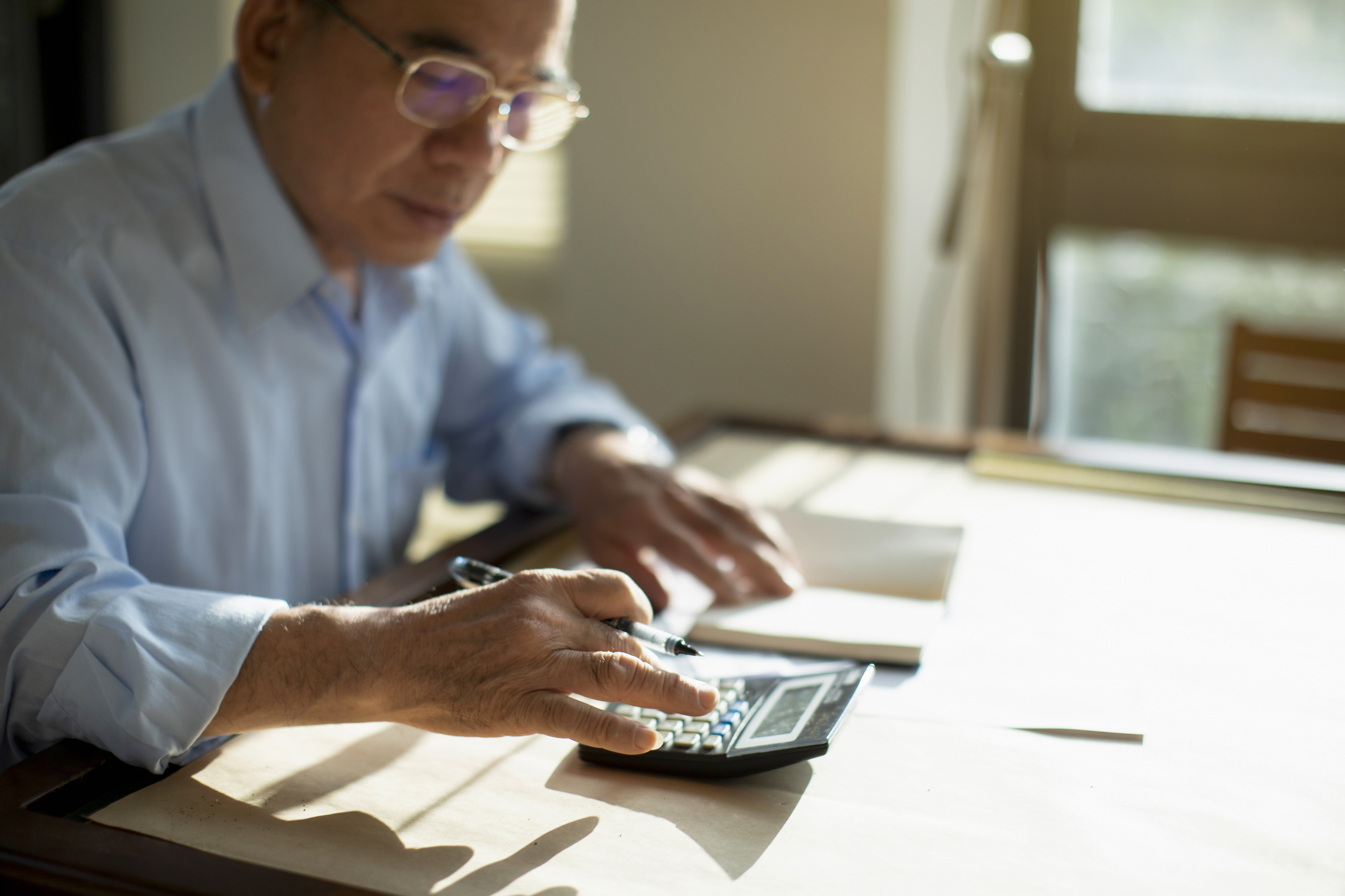 A man sits at his desk which is covered in pieces of paper. He is inputting something on a calculator.