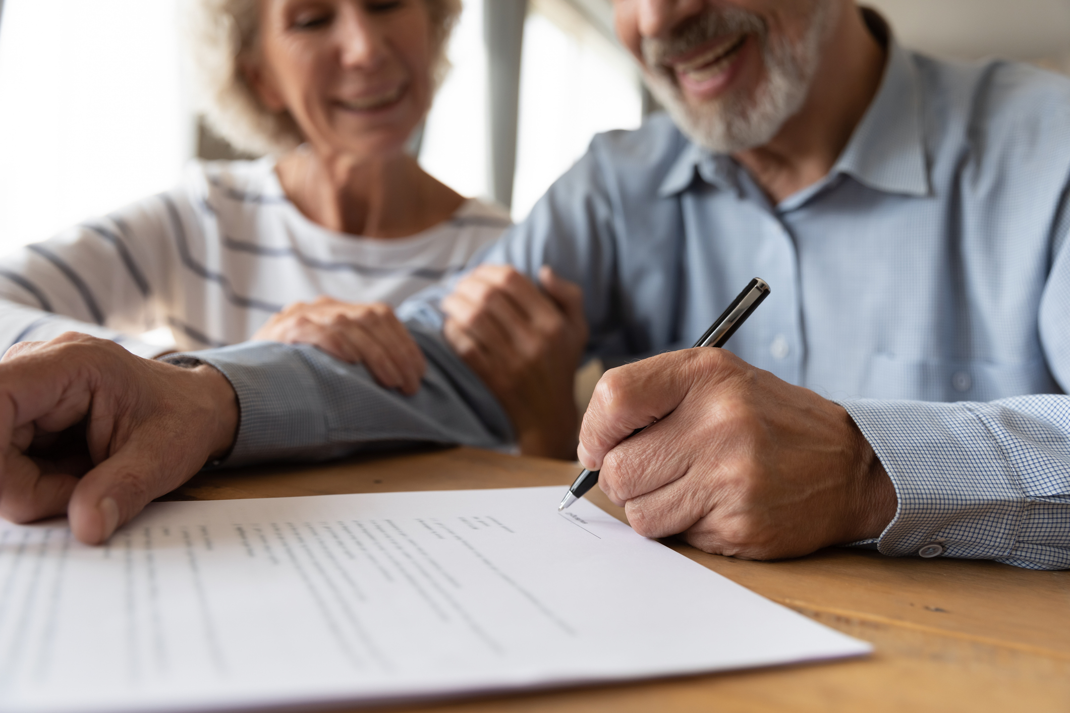 An older couple sit at a table, the woman wraps her hands around the man's arm who is signing a paper will on the table. They are both smiling.