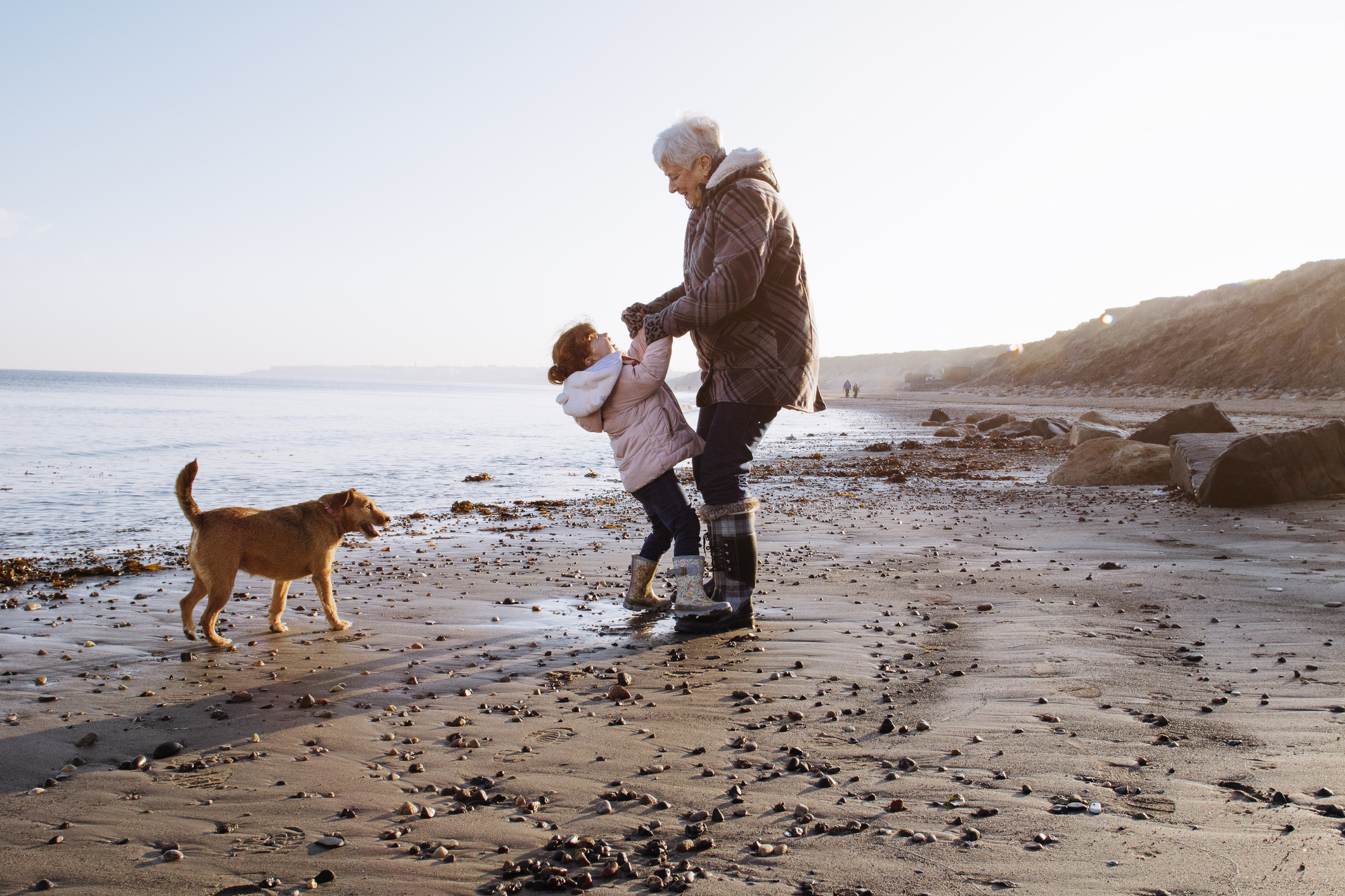 A grandma plays with her granddaughter on the beach. They are both smiling and a small dog stands off to the side watching.